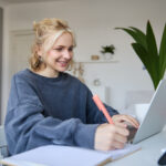 Portrait of young blond smiling woman, studying at home, remote education concept, connects to online course or lesson, writing in notebook online angol tanulas