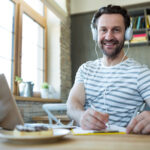 Smiling man with headphones writing in his diary at coffee shop online angol tanulas